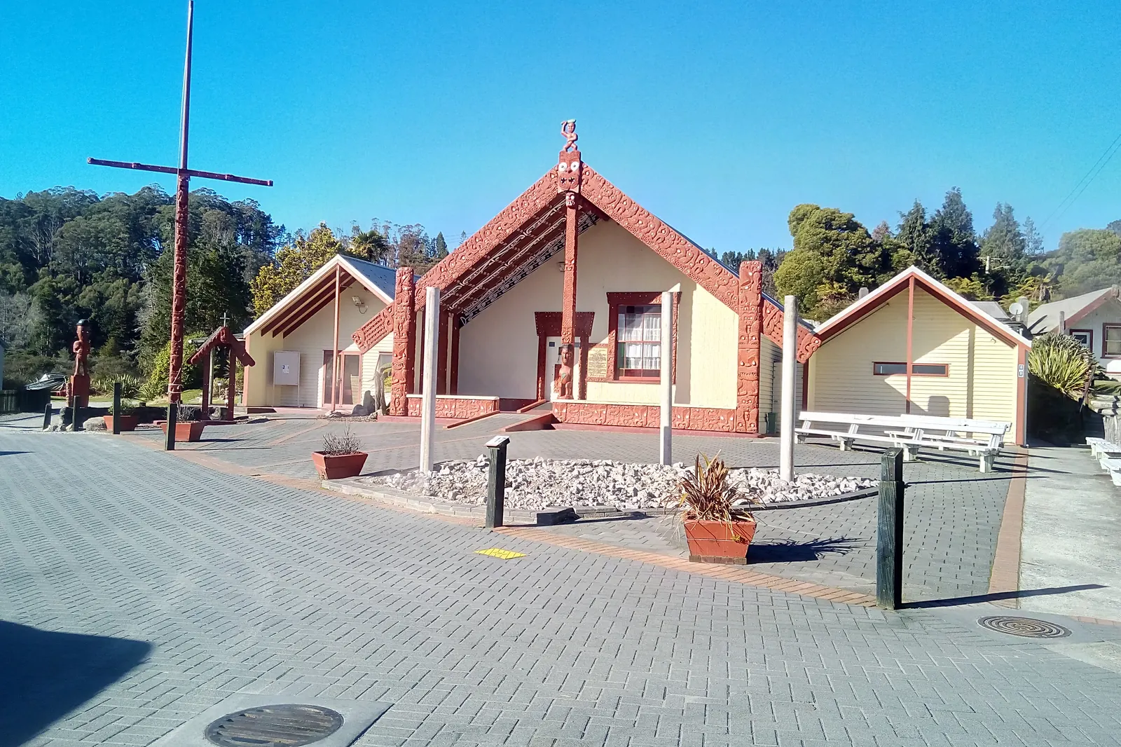 Carved and painted meeting houses with red and cream gables face a paved square in Whakarewarewa village under a blue sky