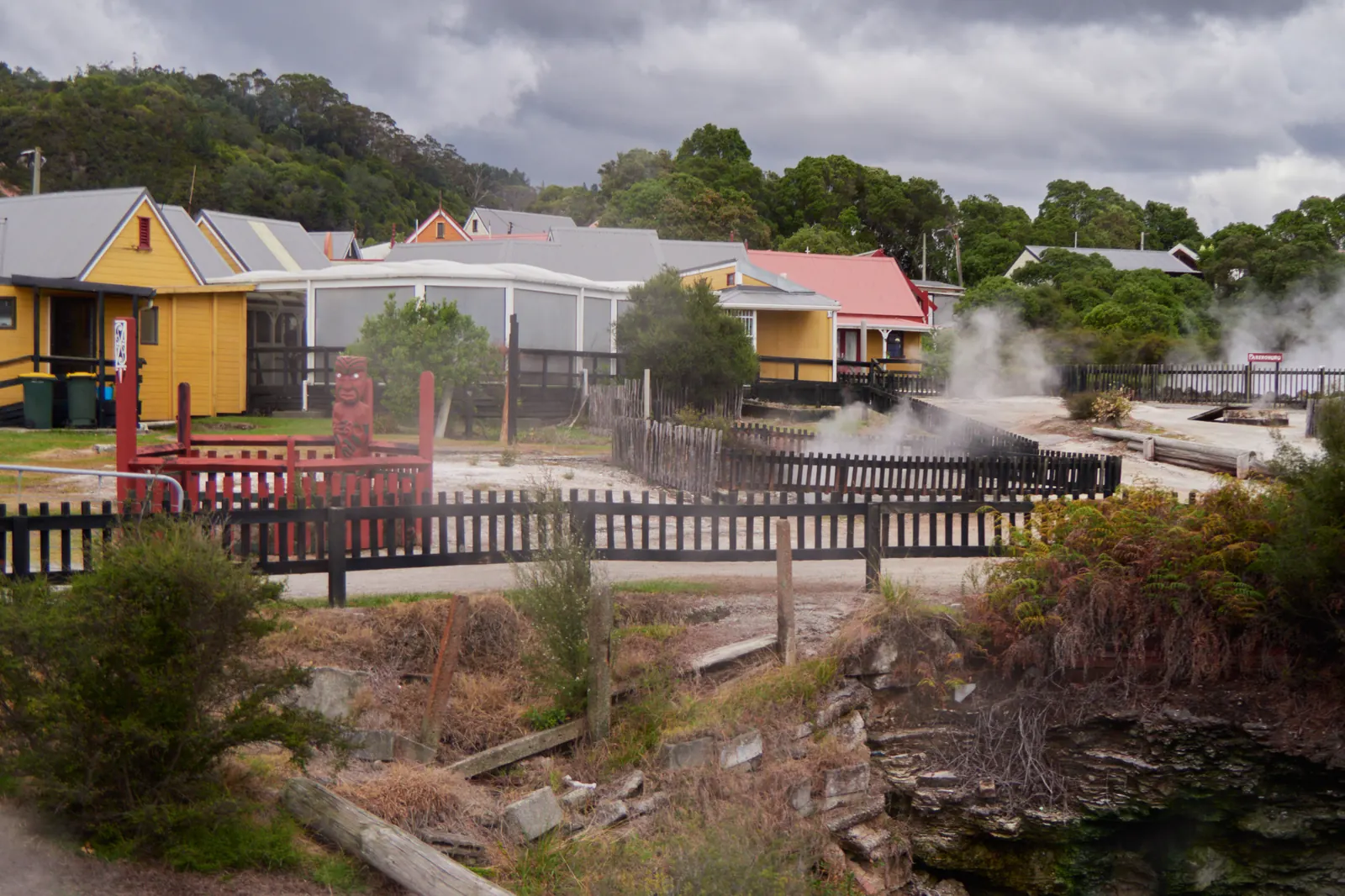 Yellow wooden houses and a red enclosure with a carved figure stand beside fenced steaming ground in Whakarewarewa village under a heavy grey sky